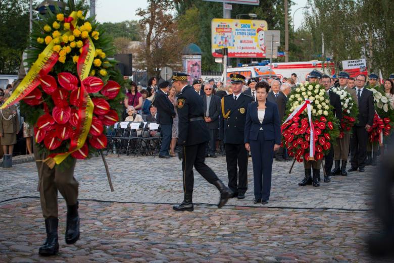 zdjęcie: na pierwszym planie widać mężczyzn w mundurach składających wiązanki z kwiatów, w tle kolejka osób z wieńcami