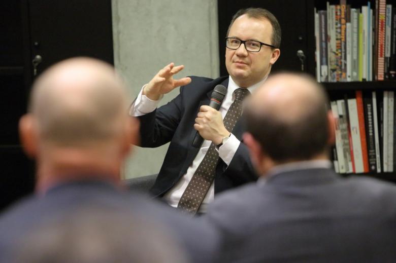 zdjęcie: na pierwszym planie głowy dwóch mężczyzn, w tle mężczyzna w garniturze, który gestykuluje