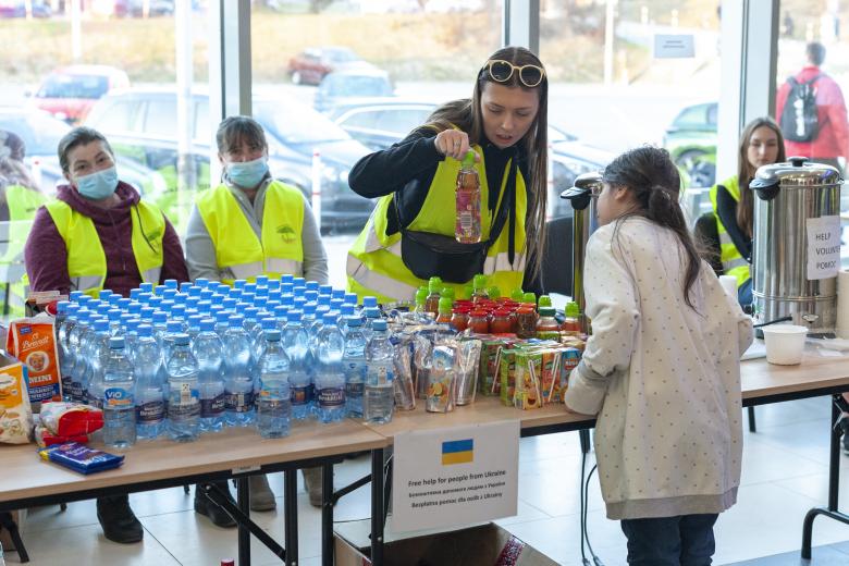 Przeszkolne pomieszczenie, w którym stoją zestawione ze sobą stoły szkolne, na których znajdują się produkty spożywcze. Stojąca przy stole wolontariuszka pokazuje sok dziecku stojącemu z drugiej strony stołu, które przygląda się mu z zainteresowaniem. Na sytuację patrzą dwie inne wolontariuszki siedzące na ławce na drugim planie.