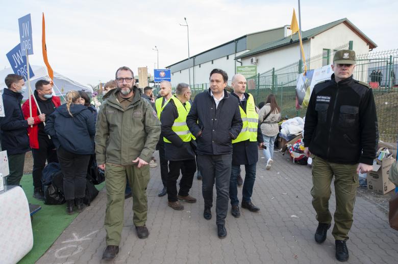 Pracownicy Biura RPO idą w towarzystwie funkcjonariuszy Straży Granicznej, rozglądając się po terenie przejścia granicznego. W tle stoi grupa ludzi trzymająca tabliczki służące do zbiórki z innymi osobami.