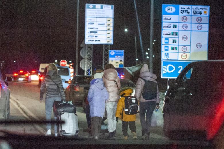 Grupa kilku osób z dziećmi, z torbami i plecakami, idzie nocą między samochodami. Na drugim planie znaki drogowe informujące o wjeździe do Polski