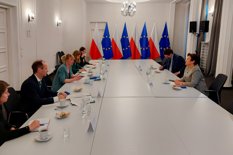 Ludzie siedzący przy długim stole, w tle flagi Polski i Unii Europejskiej