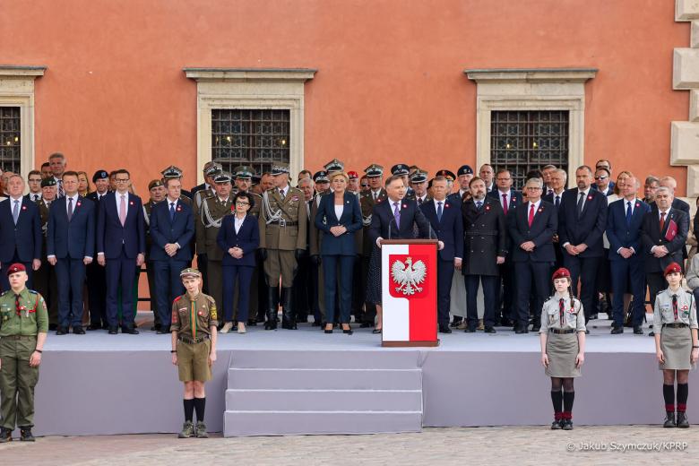 Podium, na którym znajdują się uczestnicy uroczystości trzeciomajowych. W centralnej części podium znajduje się biało-czerwona mównica z godłem Polski. Poniżej podium wartę pełnią umundurowani harcerze. Za podium znajduje się elewacja Zamku Królewskiego w Warszawie. Zdjęcie jest podpisane "Jakub Szymczuk/KPRP".