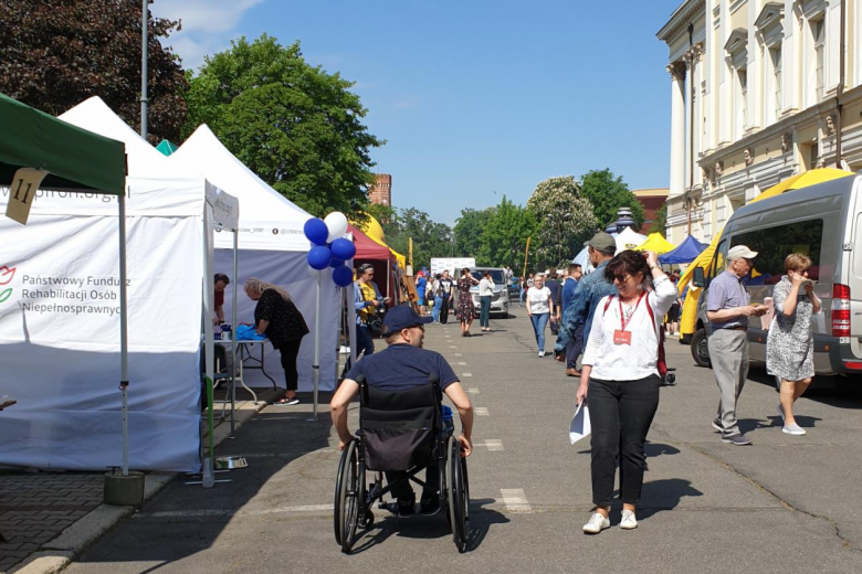 Rząd namiotów instytucji wspierających osoby z niepełnosprawnościami na ulicy, na pierwszym planie osoba na wózku