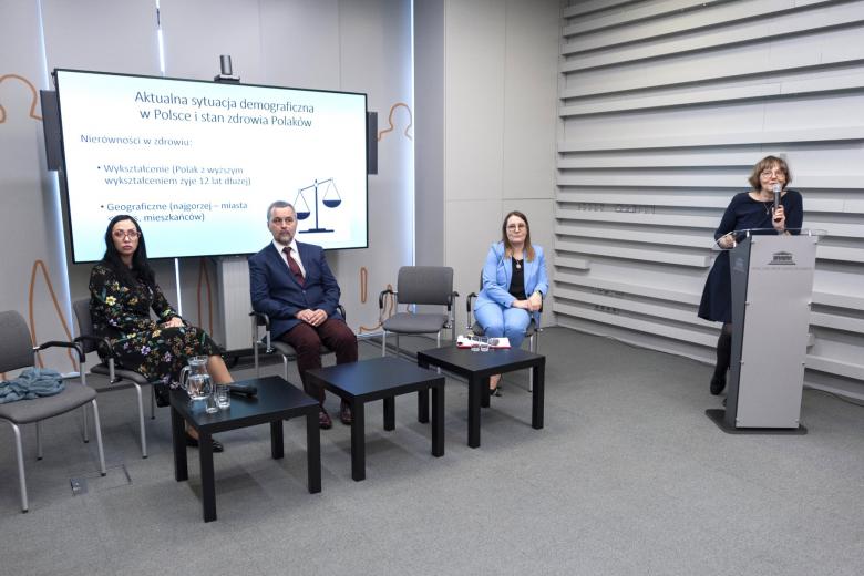 Jedna osoba przemawia, a trzy osoby siedzą za stołem w trakcie konferencji