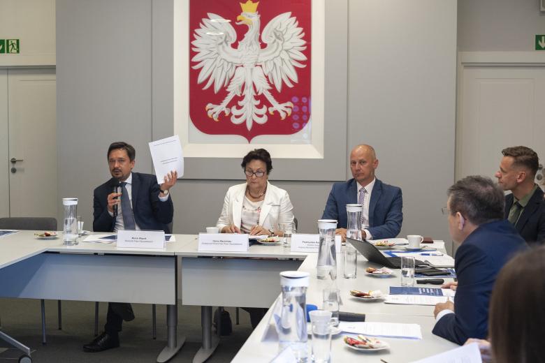 Grupa ludzi siedzących przy konferencyjnych stołach, mężczyzna przemawia do mikrofonu, w uniesionej ręce trzyma broszurę