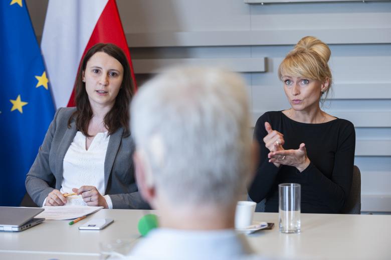 Zbliżenie na siedzących członków Komisji. Jedna z członkiń przemawia do członka komisji obróconego tyłem do kadru, a siedząca obok niej kobieta tłumaczy mowę na język migowy. W tle flagi Polski i Unii Europejskiej