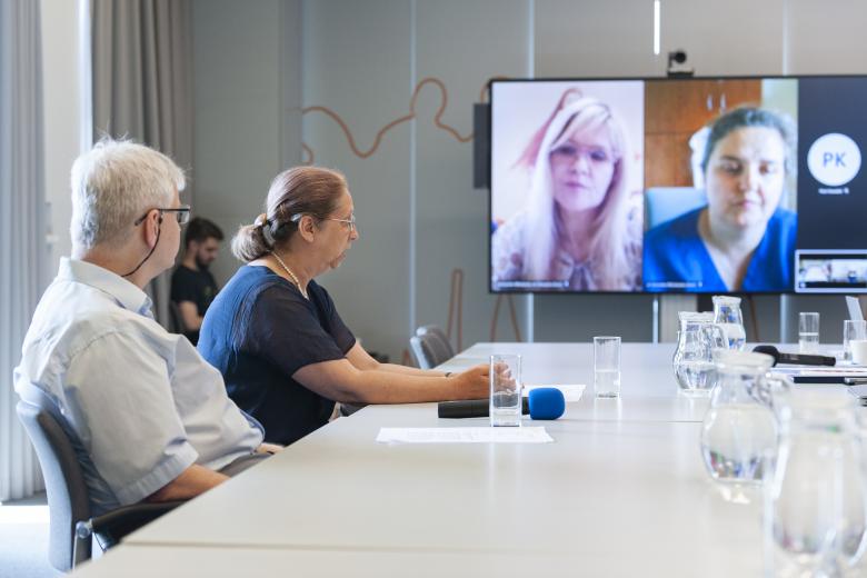 Dwóch członków Komisji siedzi przy konferencyjnym stole i jest zwróconych w stronę telewizora, na którym wyświetleni są połączeni zdalnie członkowie Komisji.