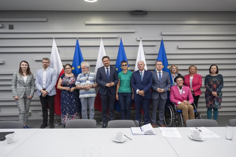 Grupa kilkunastu osób pozująca do zdjęcia na tle flag Polski i UE. W centralnym punkcie RPO Marcin Wiącek