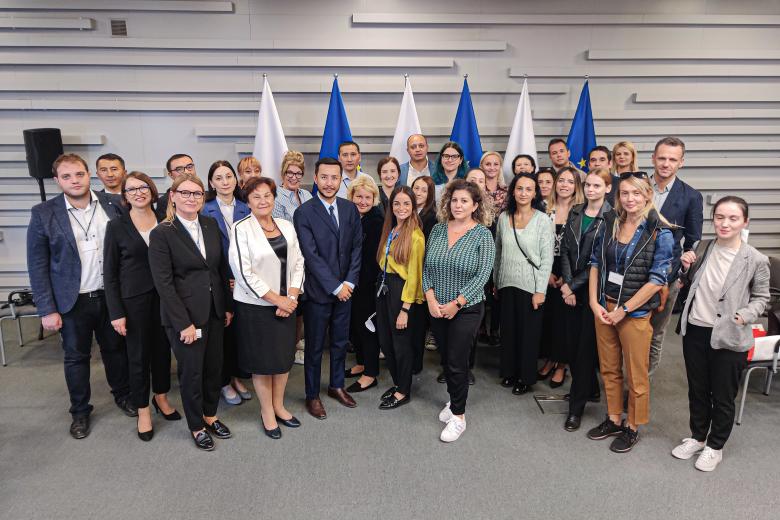 Grupa kilkudziesięciu osób pozuje do zdjęcia w pomieszczeniu na tle flag Polski i UE