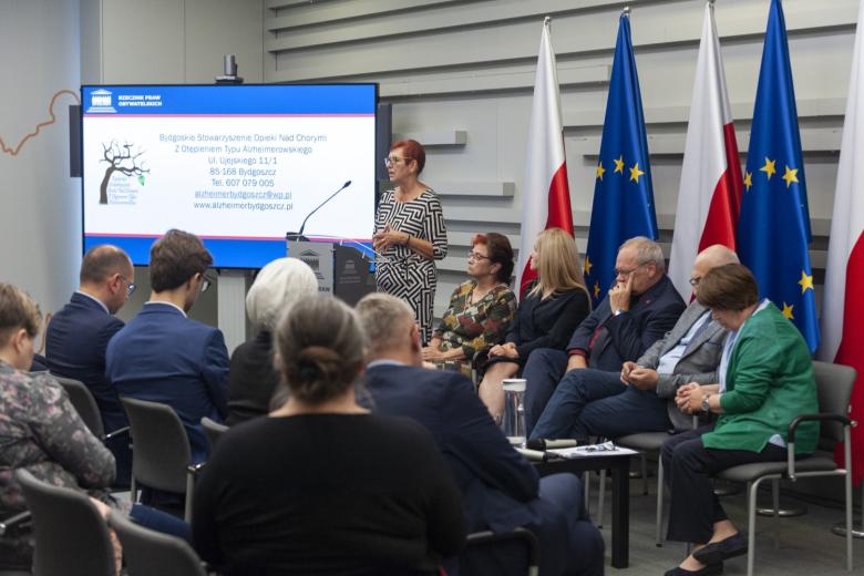 Grupa kilkudziesięciu osób siedzących na sali konferencyjnej i słuchających wypowiedzi prelegentki stojącej za mównicą