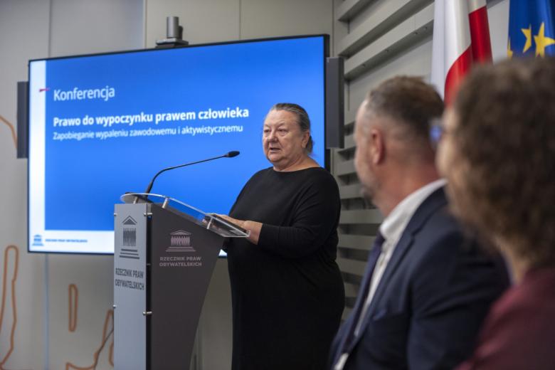 Dyrektorka Centrum Projektów Społecznych Barbara Imiołczyk przemawia za mównicą, w tle duży ekran z napisem "Prawo do wypoczynku prawem człowieka. Zapobieganie wypaleniu zawodowemu i aktywistycznemu" 