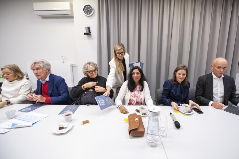 Goście siedzą przy długim stole konferencyjnym. Stojąca nad nimi osoba rozdaje im egzemplarze publikacji.