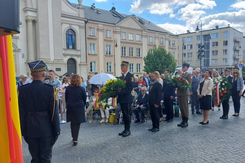 Osoby, w tym ZRPO Hanna Machińska, stoją w oczekiwaniu na złożenie wieńców pod tablicą. 