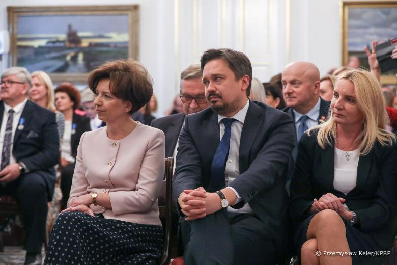 RPO Marcin Wiącek siedzi w pierwszym rzędzie krzeseł w dużej zabytkowej sali. Na drugim planie kilkadziesiąt osób siedzi w dalszych rzędach