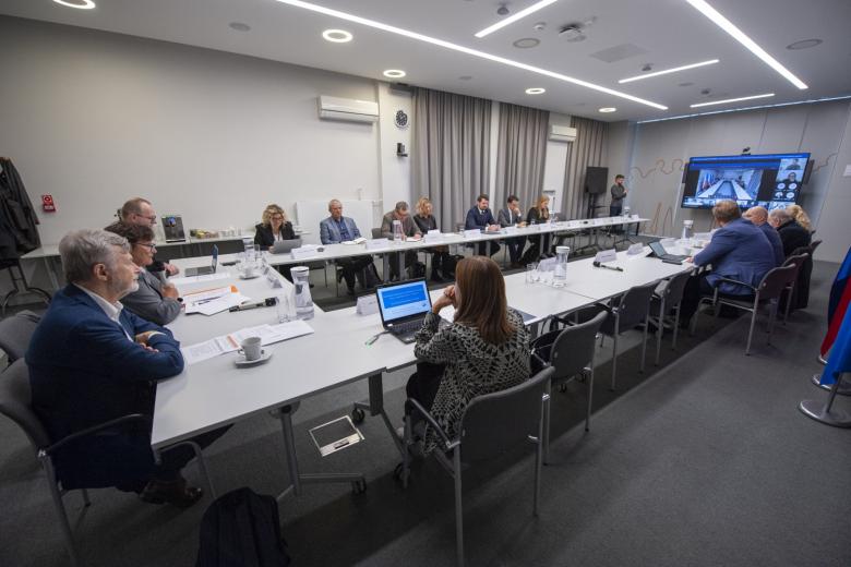 Grupa kilkunastu osób siedzących przy stole konferencyjnym, w tle duży ekran telewizora z obrazem osób uczestniczących w spotkaniu zdalnie