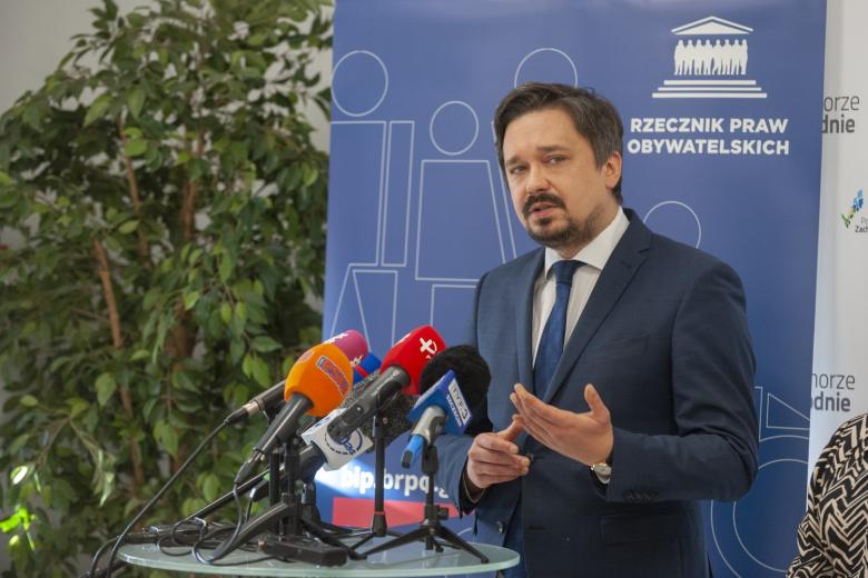 RPO Marcin Wiącek stoi na tle planszy z napisem "Rzecznik Praw Obywatelskich" i mówi do kilku mikrofonów stojących przed nim na stole