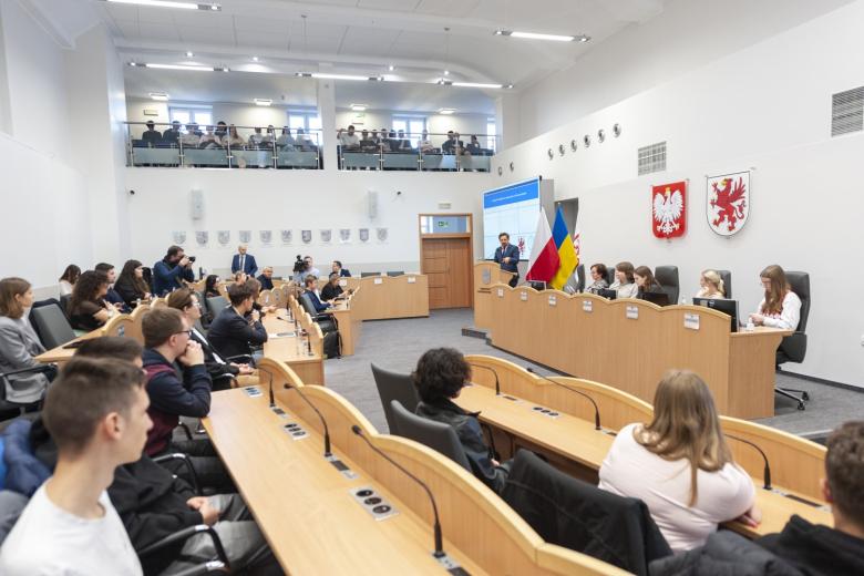 Duża sala posiedzeń, w której siedzą młodzi ludzie i słuchają wystąpienia osoby stojącej za mównicą, w oddali na balkonie również słuchające osoby, na ścianie herb Polski i województwa zachodnipomorskiego