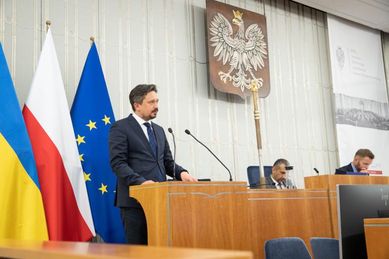 RPO Marcin Wiącek przemawia z podium w sali plenarnej Senatu. Na drugim planie, w miejscach prezydium, siedzą dwie osoby. W tle godło Polski oraz flagi Polski, Ukrainy oraz Unii Europejskiej