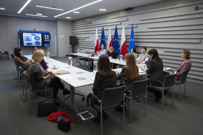 Uczestnicy warsztatu słuchają prezentacji wyświetlanej na telewizorze konferencyjnym. W tle flagi Polski i Unii Europejskiej