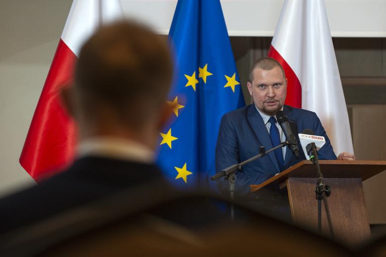 Tomasz Przybysz-Przybyszewski wypowiada się z podium, spoglądając w stronę laureata, którego tył sylwetki jest na pierwszym planie. W tle flagi Polski i Unii Europejskiej