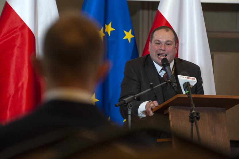 Michał Piechel wypowiada się z podium spoglądając w stronę laureata, którego tył sylwetki jest na pierwszym planie. W tle flagi Polski i Unii Europejskiej