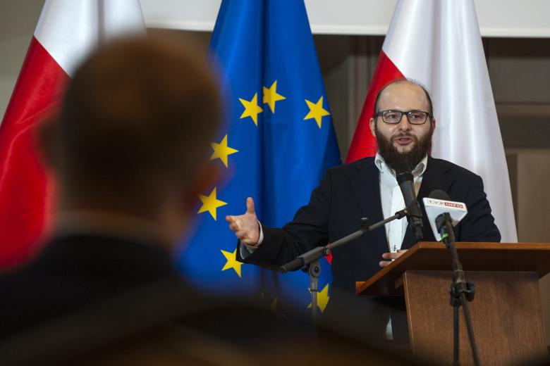 Adam Zawisny wypowiada się z podium w stronę laureata, gestykulując prawą dłonią. Tył sylwetki laureata widać na pierwszym planie. W tle flagi Polski i Unii Europejskiej