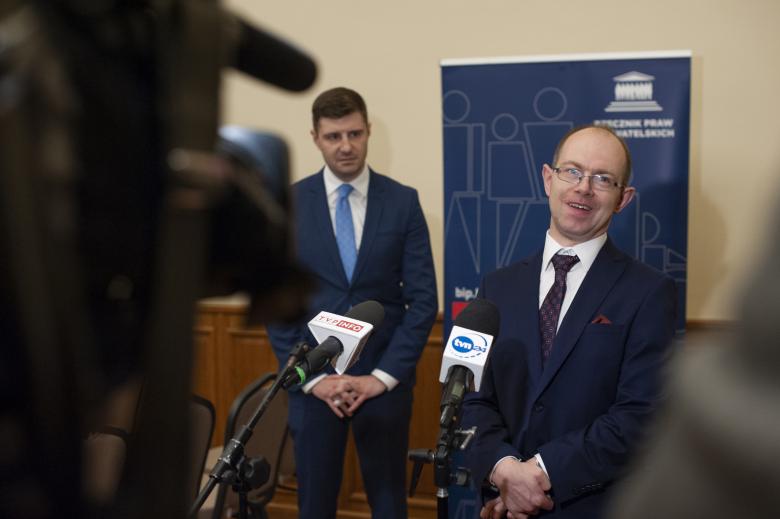 Krzysztof Kurowski wypowiada się do dwóch mikrofonów medialnych. Na pierwszym planie po lewej stronie jest kamera telewizyjna. Na drugim planie stoi przyglądający się Krzysztofowi Kurowskiemu Valeri Vachev. W tle stoi baner Biura RPO. 