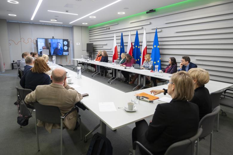 Członkowie Komisji siedzą przy podkowiastym stole w sali konferencyjnej i patrzą w stronę telewizora konferencyjnego, na którym wyświetlany jest przemawiający członek Komisji.