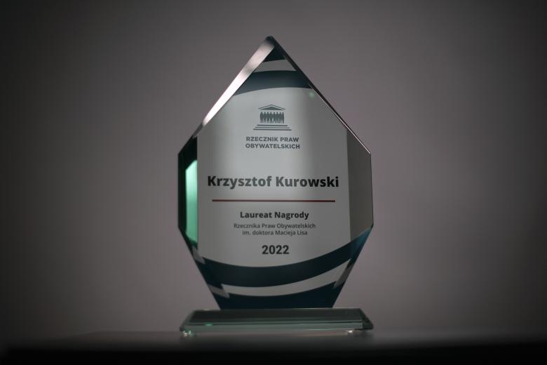 Szklana statuetka o kształcie siedmiokątu z logiem RPO i napisem "Krzysztof Kurowski - Laureat Nagrody Rzecznika Praw Obywatelskich im. doktora Macieja Lisa - 2022"