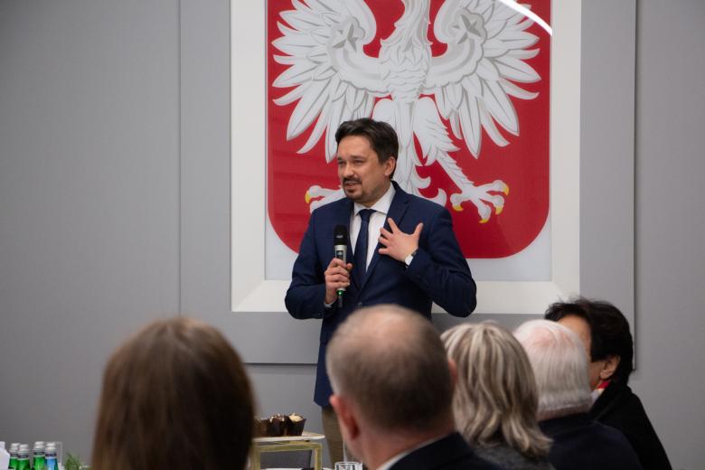 RPO Marcin Wiącek wypowiada się w stronę siedzących za stołem gości. Na pierwszym planie widoczne od tyłu sylwetki pięciu osób. W tle godło Polski