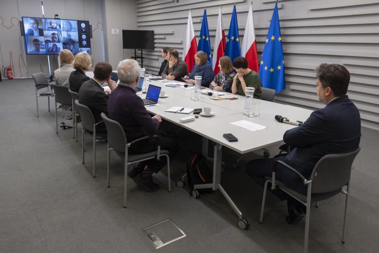 Grupa kilku osób siedząca po obu stronach dużego stołu konferencyjnego, w oddali duży ekran z widocznym ekranem z uczestnikami spotkania w aplikacji do komunikowania się na odległość