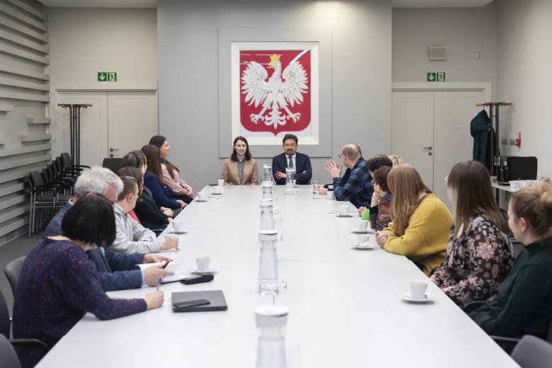 Grupa kilkunastu osób siedzi przy dużym stole konferencyjnym, w tle godło Polski