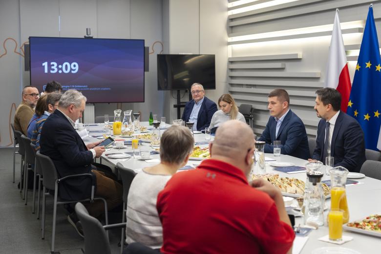 RPO Marcin Wiącek, ZRPO Wojciech Brzozowski oraz dziennikarze rozmawiają przy stole w sali konferencyjnej. Za Rzecznikiem stoją flagi Polski i Unii Europejskiej