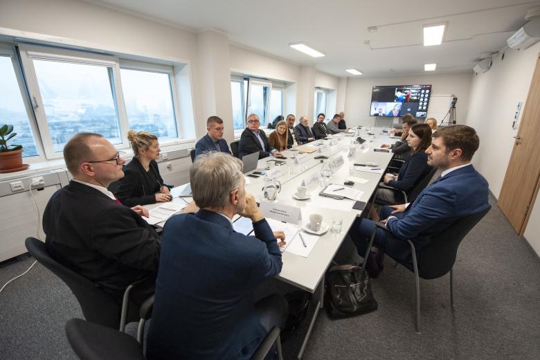 Członkowie Komisji dyskutują przy długim konferencyjnym stole. U szczytu stoły znajduje się ekran wyświetlający uczestników łączących się za pomocą Intenetu. 