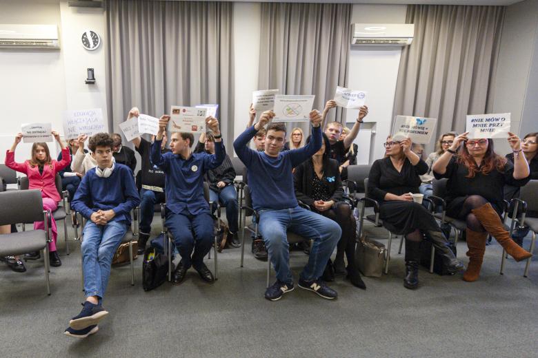 Uczestnicy spotkania siedząc podnoszą do góry kartki z postulatami dotyczącymi walki z wykluczeniem osób w spektrum autyzmu.