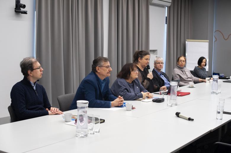 Członkowie komisji dyskutują przy długim stole konferencyjnym. Kadr zwrócony jest na jedną ze stron stołu, przy której siedzi siedem osób. Kobieta siedząca w środku trzyma mikrofon i wypowiada się do niego, reszta osób słucha.