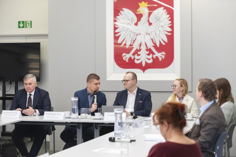 Członkowie Komisji Ekspertów siedzą przy konferencyjnym stole. W centrum stołu siedzą ZRPO Wojciech Brzozowski (który wypowiada się do mikrofonu) i dyrektor Piotr Mierzejewski. W tle godło Polski