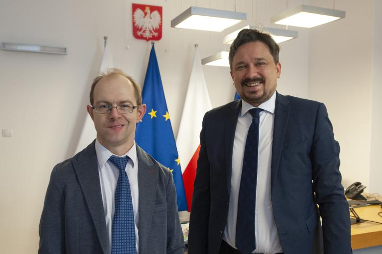 Marcin Wiącek i Krzysztof Kurowski pozują do zdjęcia w gabinecie RPO. W tle godło Polski oraz flagi Polski i Unii Europejskiej