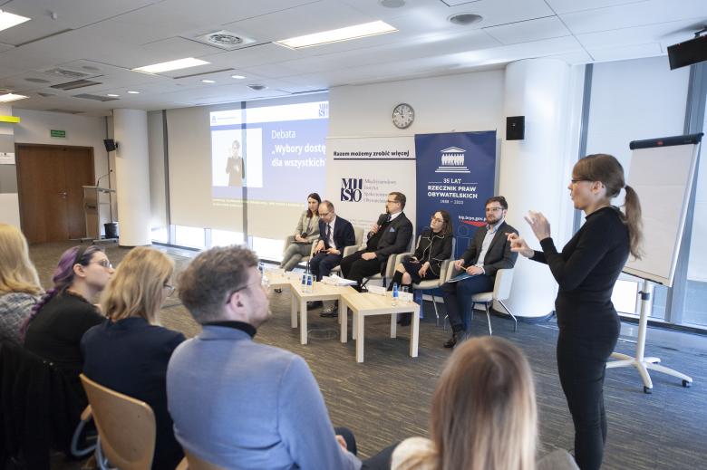 Członkowie panelu rozmawiają siedząc w fotelach na scenie. Na lewo od nich pracuje tłumaczka języka migowego.