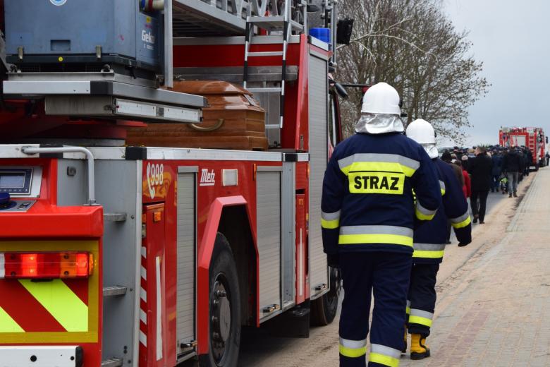 Orszak idący drogą, w orszaku strażacy w mundurach oraz wóz bojowy straży pożarnej, na którym leży trumna