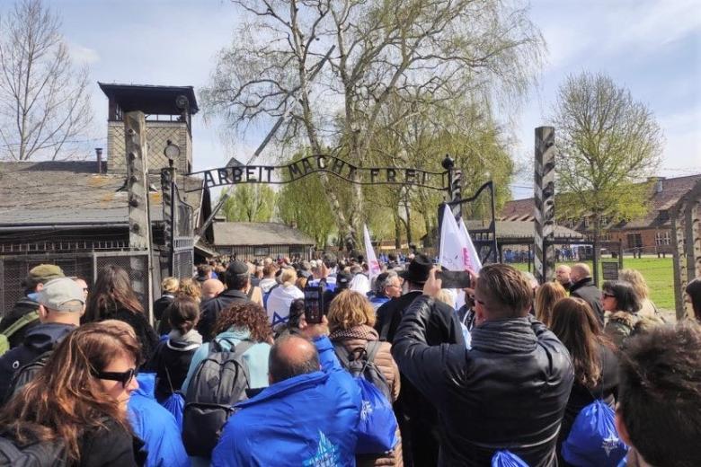 Uczestnicy marszu przechodzą pod bramą obozu z napisem "Arbeit Macht Frei". Niektórzy z nich są ubrani na niebiesko i niosą flagi Izraela