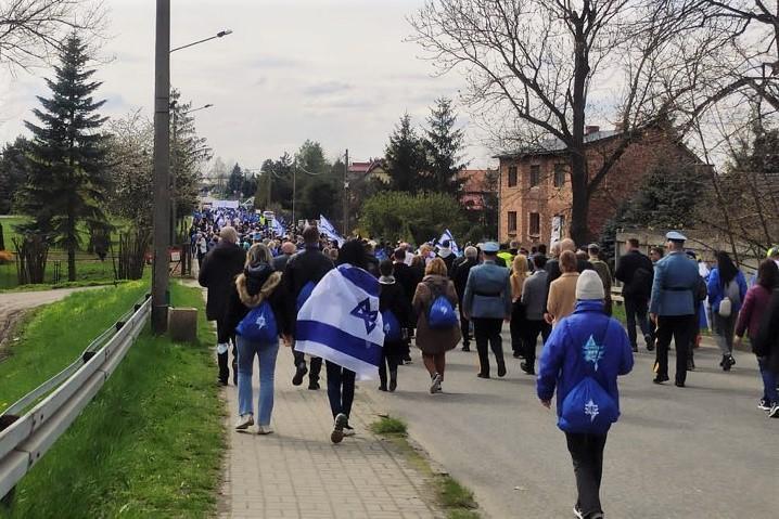 Kilkaset osób, niektórzy niosą biało niebieskie flagi idzie asfaltową drogą