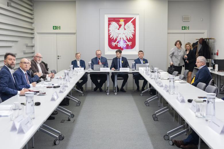 Członkowie Komisji siedzą przy stole. W szczycie stołu siedzi dwóch przewodniczących Komisji oraz zastępcy RPO Valeri Vachev i Wojciech Brzozowski. W tle godło Polski.