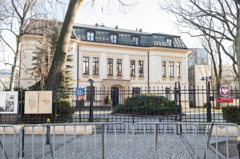 Budynek Trybunału Konstytucyjnego w Warszawie. Przed budynkiem jest metalowe ogrodzenie z bramą, przy której wisi tabliczka z napisem "Trybunał Konstytucyjny". Kilka metrów przed płotem stoją metalowe, tymczasowe bariery.