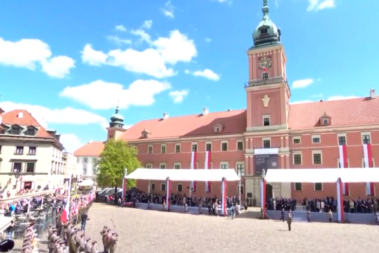 Widok ogólny Placu Zamkowego w Warszawie przygotowanego na uroczystości państwowe. Centralną część kadru zajmuje zamek przyozdobiony biało-czerwonymi flagami. Przed zamkiem stoją trybuny dla gości. Po drugiej stronie placu stoją widzowie oraz żołnierze w ceremonialnych mundurach.