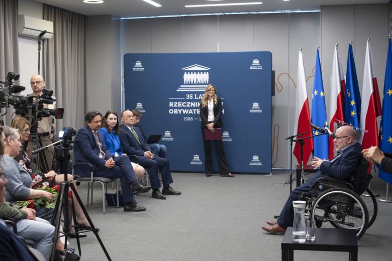 Jan Filip Libicki siedzący na wózku i przemawiający do mikrofonów na tle flag Polski i UE