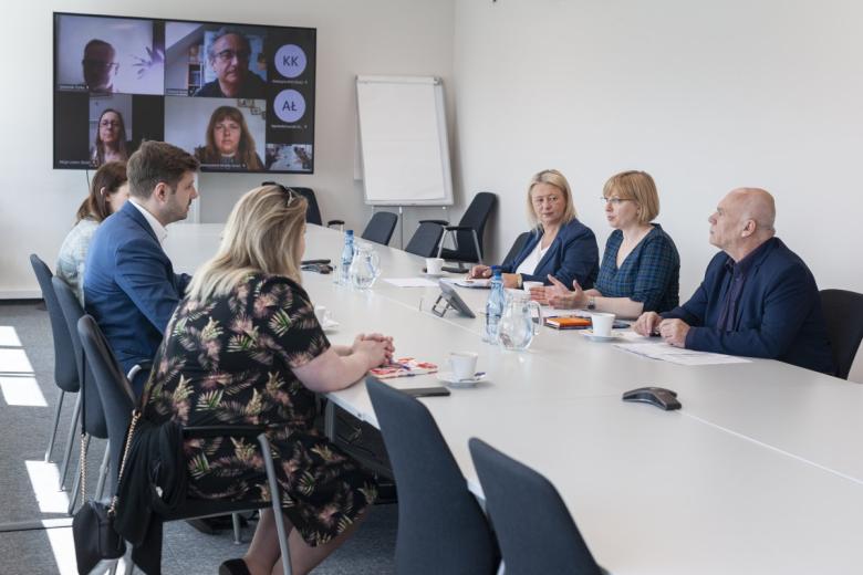 Sześć osób siedzi po dwóch stronach długiego, prostokątnego stołu, na długim boku duży ekran z uczestnikami spotkania łączącymi się za pomocą programu do komunikowania się na odległość