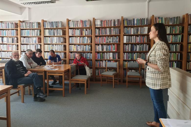 W sali biblioteki z półkami pełnymi książek osoba przemawia na stojąca do siedzących przy stołach osób