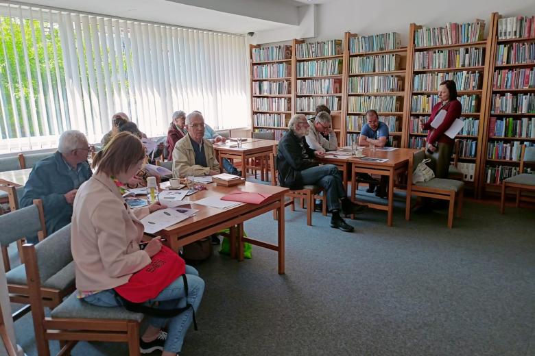 Kilkanaście osób siedzi przy stołach w sali biblioteki. Przy ścianach półki pełne książek.